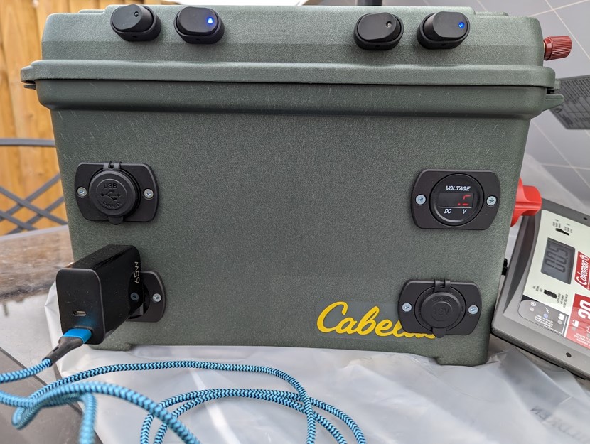 Olive-green portable battery box with a red master switch, digital voltmeter, USB ports, inverter outlets, and Anderson Powerpoles