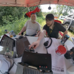 Two operators at a Field Day station under a red canopy with radios, laptop, and logging gear