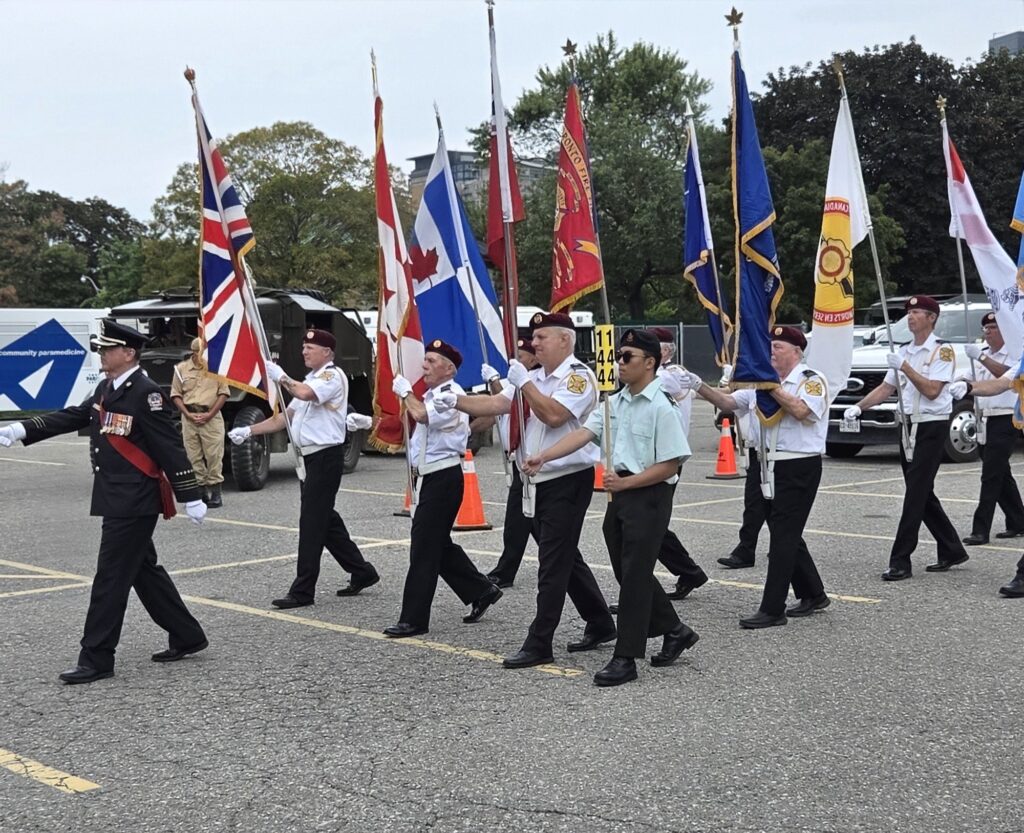 Colour guard marching with multiple flags including the Canadian Maple Leaf, the Union Jack, and a Toronto Fire Services flag, led by an officer in dress uniform with medals and a red sash.