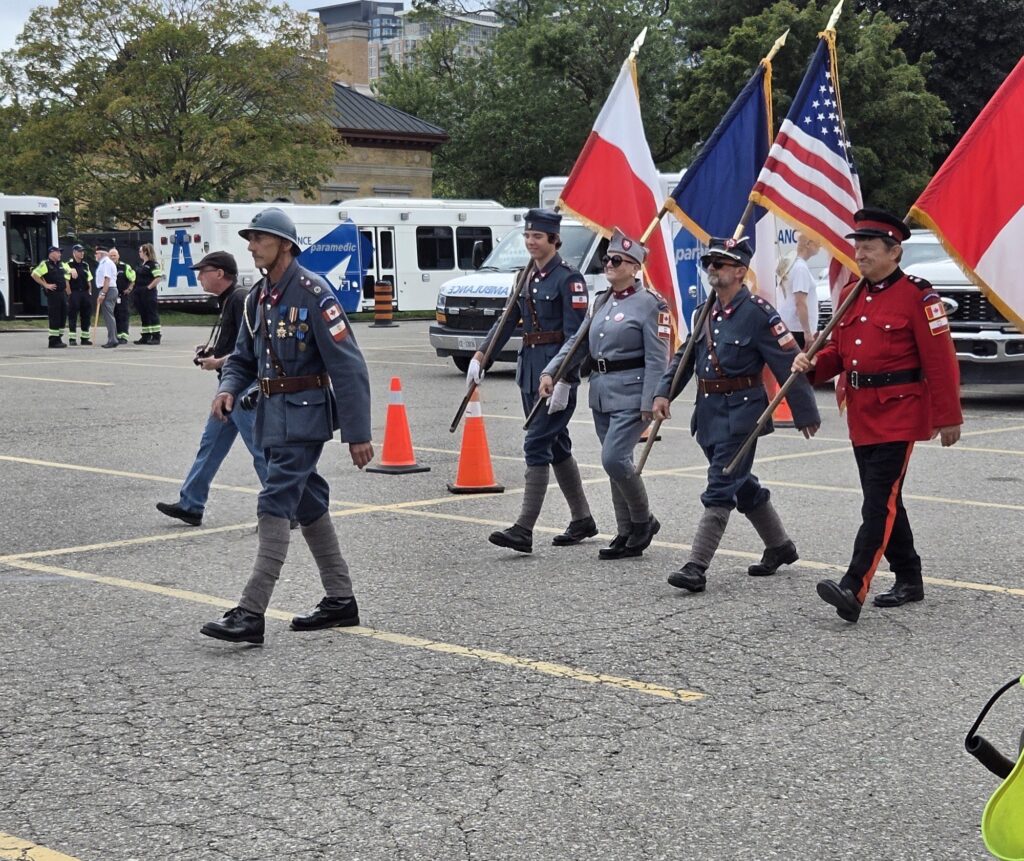 Colour party in historical military uniforms — including a WWI-style Adrian helmet and a bright red dress tunic — marching with flags including the United States flag, with paramedic vehicles in the background.
