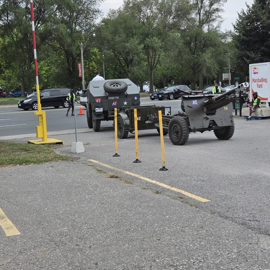 Vintage olive-drab military truck towing a field artillery cannon at the CNE Marshalling Yard during Warriors’ Day, with white star markings and event staff in a high-visibility vest nearby.