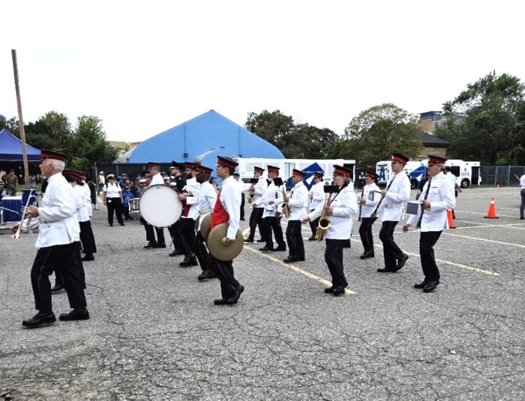 Marching band in white tunics, black trousers, and red-banded caps performing with brass, woodwind, and percussion instruments on a paved outdoor staging area during Warriors’ Day.
