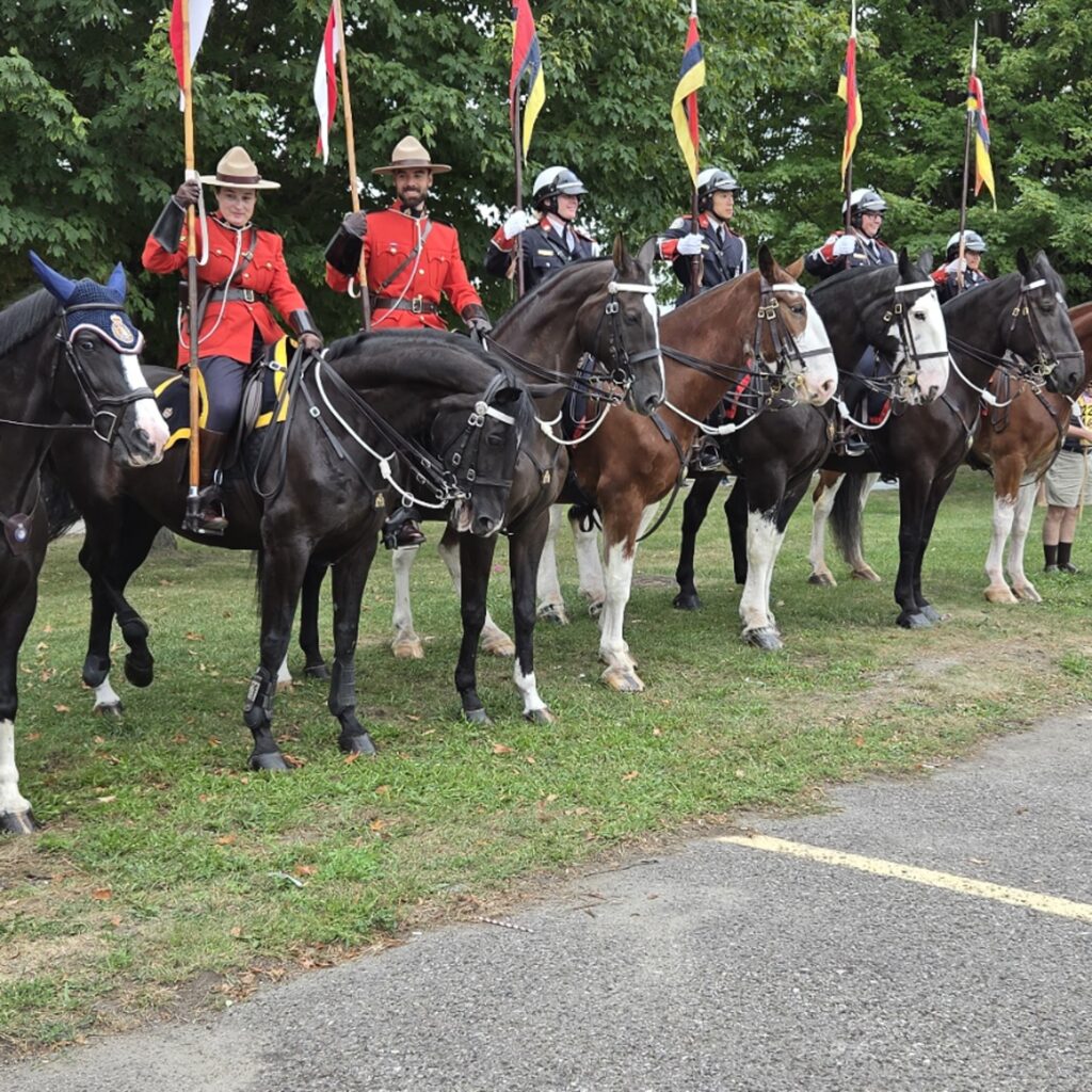 Ceremonial mounted police in formation, including RCMP officers in red serge and Stetson hats followed by riders in dark blue uniforms with silver helmets, each holding a lance with a coloured pennant.