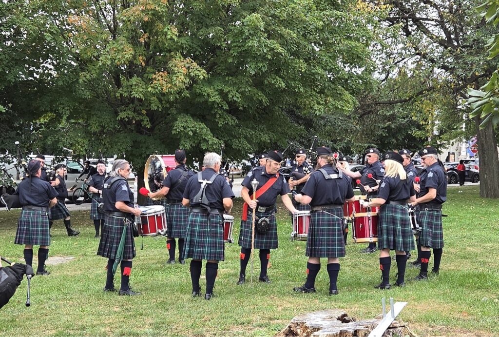 Pipe and drum band members in tartan kilts and Glengarry caps gathering on a grassy park area with bagpipes and red drums, led by a drum major in a red sash holding a mace.