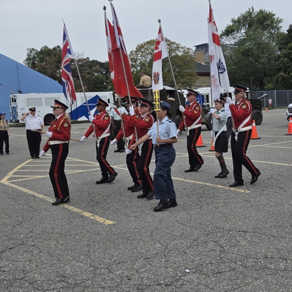 Parade participants in red ceremonial tunics and other uniforms marching with the Union Jack, Canadian Red Ensign, and Maple Leaf flags while a uniformed officer salutes nearby.