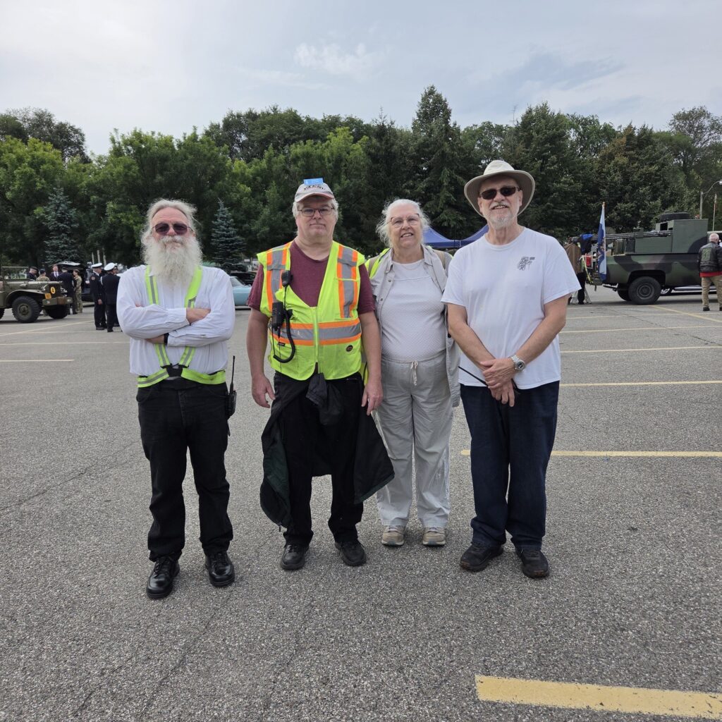 Four Skywide Amateur Radio Club volunteers at Warriors’ Day 2026 — David; Roger Hopson (VA3HPR); Candace McCool (VA3KKE); and Ken Baine (VA3KEX) — standing outdoors near military vehicles, with two wearing high-visibility safety vests and radios.