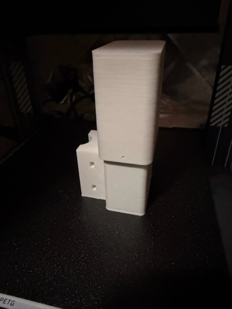 Printed white lid stacked on the enclosure body on a dark printer bed. The side saddle has two mounting holes. The bed is labeled PETG.