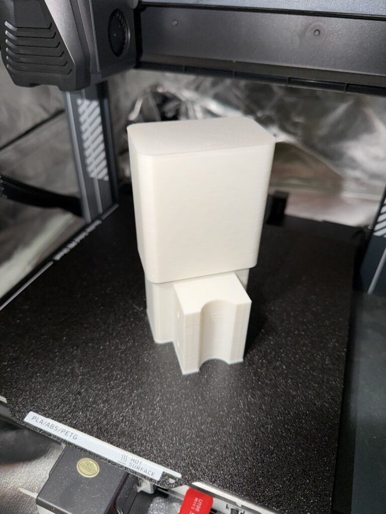Closed white 3D-printed enclosure standing on the printer bed, lid on, pole saddle facing the camera.