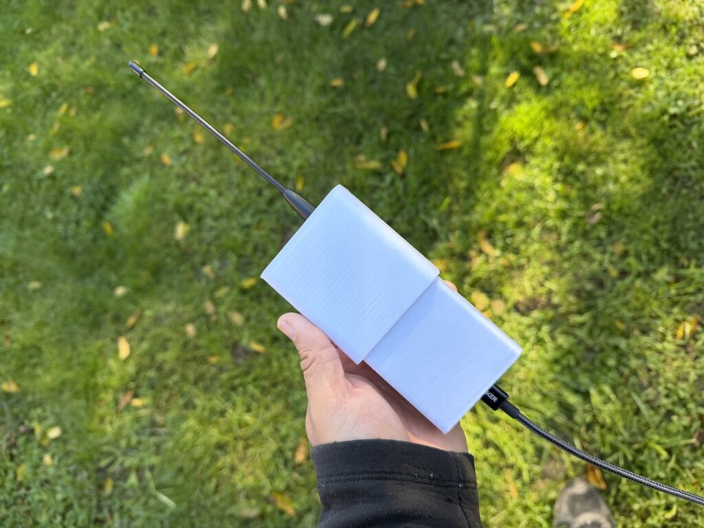Hand holding a white 3D-printed enclosure over grass, whip antenna on top and a braided power cable out the bottom.