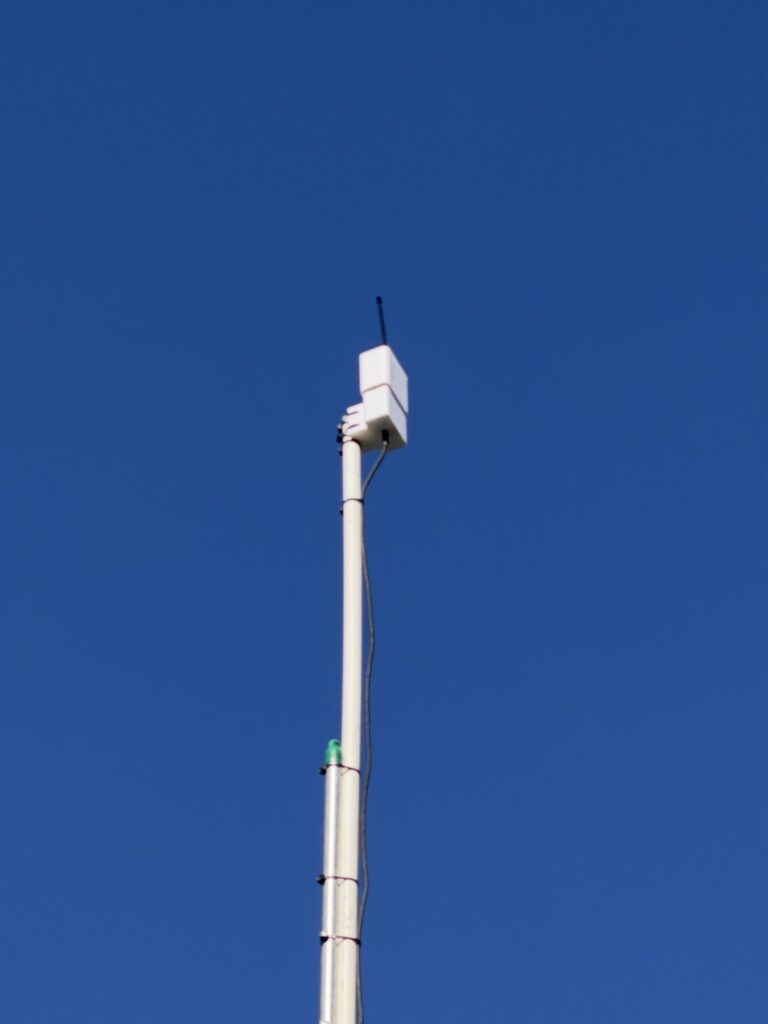 Close view of the white printed box mounted at the top of the mast, antenna pointing up, with the feed cable tied down the pole.