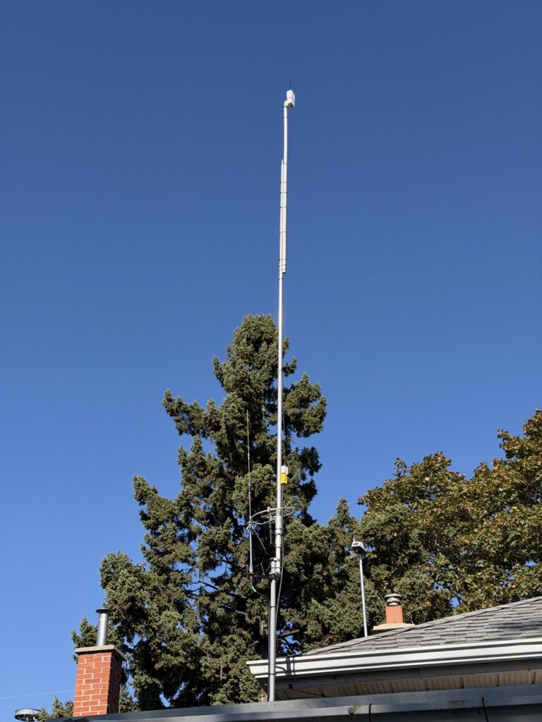 White enclosure at the top of a tall mast above a house roof and brick chimney, against a clear blue sky and evergreen trees.