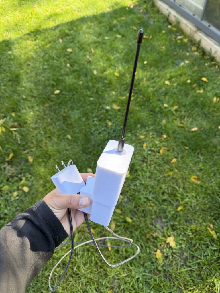 Hand holding the finished white enclosure with whip antenna, pole saddle, and a white wall-wart power adapter on the cable.