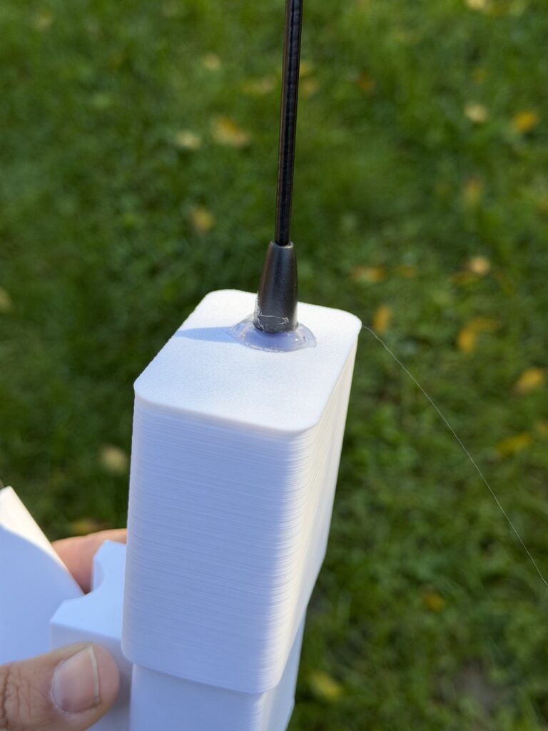 Close-up of the black whip antenna at the top of the white printed box, sealed with a ring of clear silicone.